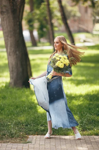Güneşli bir bahar parkında elbise giymiş şık bir kız. Güzel bir kızın sakin portresi. Baharda elinde bir buketle duruyor. — Stok fotoğraf