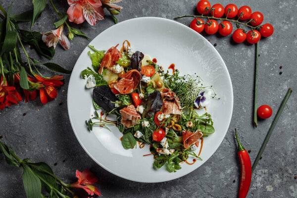 salad with ham and pear on dark gray background. jamon salad, salad mix, spinach, pear, cherry tomatoes, Dor Blue, capers and vinegar dressing Bianco