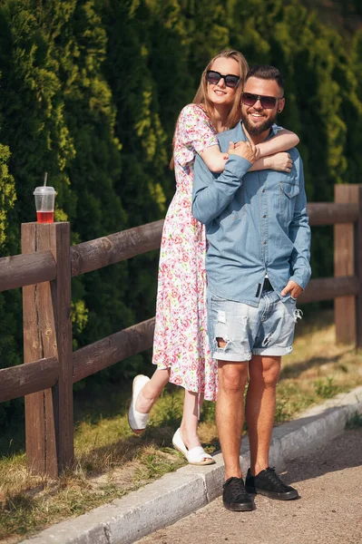 Uma menina bonita e macia com cabelo loiro, um vestido leve e um buquê caminha em um parque ensolarado com seu namorado bonito em uma camisa azul e shorts. Dia ensolarado. Verão — Fotografia de Stock