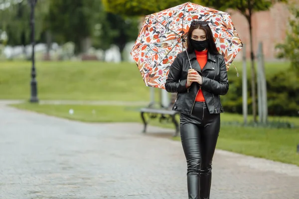 A masked girl is walking along the street. A girl in a protective mask walks in the park with an umbrella in the rain. Coronavirus infection COVID-19.