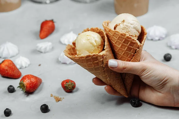 ice cream in a waffle cone on a light background in a hand. The ice cream is melting. Waffle cones and strawberries in the background