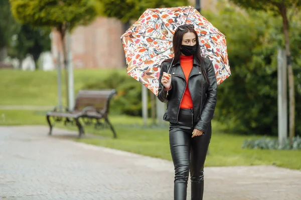 Une fille masquée marche dans la rue. Une fille portant un masque de protection se promène dans le parc avec un parapluie sous la pluie. Infection à coronavirus COVID-19 — Photo
