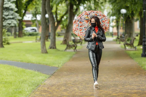 Ein maskiertes Mädchen läuft die Straße entlang. Ein Mädchen mit Schutzmaske geht mit einem Regenschirm im Regen durch den Park. Coronavirus-Infektion COVID-19 — Stockfoto