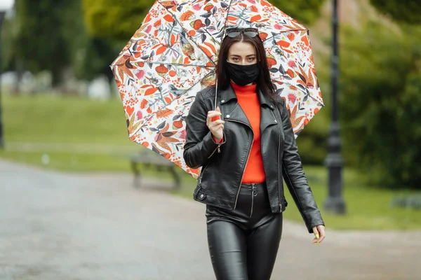 Po ulici kráčí maskovaná dívka. Dívka v ochranné masce chodí do parku s deštníkem v dešti. Koronavirová infekce COVID-19 — Stock fotografie