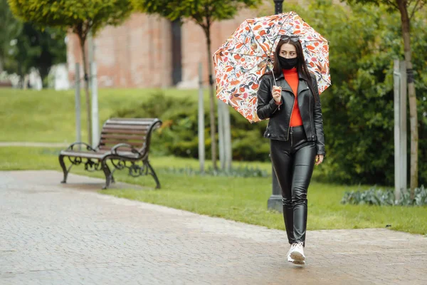 Uma menina mascarada está andando pela rua. Uma menina com uma máscara protetora caminha no parque com um guarda-chuva na chuva. Infecção pelo vírus da coronária COVID-19 — Fotografia de Stock