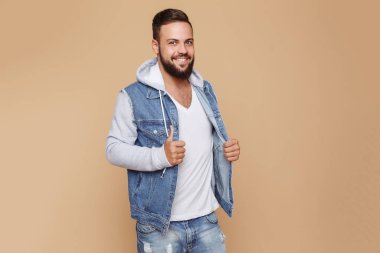 Güzel sakallı, kot ceketli ve krem rengi arka planda beyaz tişörtlü şık, genç ve neşeli bir adam. Denim ceket reklam panosu konsepti