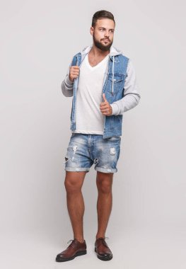 Kot ceketli, güzel sakallı, beyaz tişörtlü ve gri arka planda kot şortlu şık, genç ve neşeli bir adam. Denim ceket ve kot şort reklam panosu konsepti