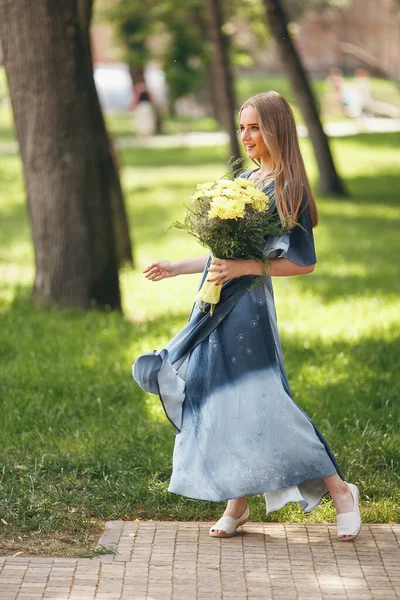 Ragazza elegante in posa in un abito in un parco primaverile soleggiato. Ritratto tranquillo di una bella ragazza in piedi con un bouquet in primavera — Foto Stock