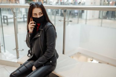 Alışveriş merkezinde siyah maskeli ve cep telefonu olan bir kız. Koronavirüs salgını. Maskeli bir kadın alışveriş merkezinde duruyor. Koruyucu maskeli bir kız alışveriş merkezinde alışveriş yapıyor..