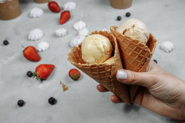 Waffle külahında dondurma, arka planda hafif bir el. Dondurma eriyor. Waffle konileri ve çilekler arka planda..