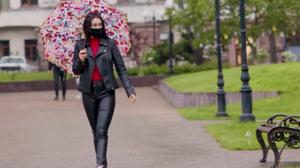 En flicka i en skyddande svart mask går genom parken med ett paraply i regnet. En maskerad flicka går längs gatan. Coronavirusinfektion COVID-19 — Stockvideo