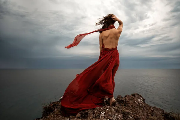 バック夕日の間に海の上の岩の上に赤い長いドレスのポーズで美しい官能的な女性を表示します 空中の暗い雲 空気中を飛ぶ布のストリップ — ストック写真