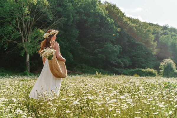 A young woman in a white sundress, a wreath of daisies with a large wicker bag on her shoulder is walking through a field of daisies, against the background of the forest, the sunset light.