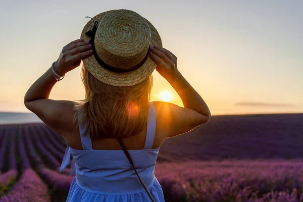 Uma linda loira está de pé em um campo de lavanda com as costas, segurando um chapéu em suas mãos e desfrutando do nascer do sol. O conceito de aromaterapia, óleo de lavanda, sessão de fotos na lavanda . — Fotografia de Stock