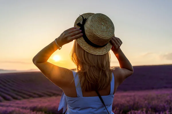 Uma linda loira está de pé em um campo de lavanda com as costas, segurando um chapéu em suas mãos e desfrutando do nascer do sol. O conceito de aromaterapia, óleo de lavanda, sessão de fotos na lavanda . — Fotografia de Stock
