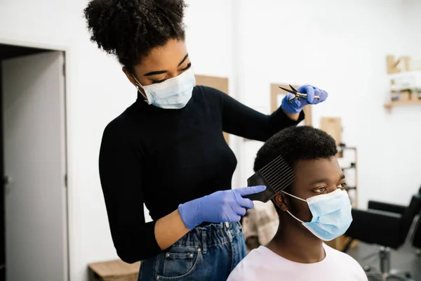 Afro-Amerikan güzellik kuaförü. Kendini koronavirüs salgınından korumak için hem maske hem de eldiven takan Afro-Amerikan bir müşteriyi soyup tarıyor.