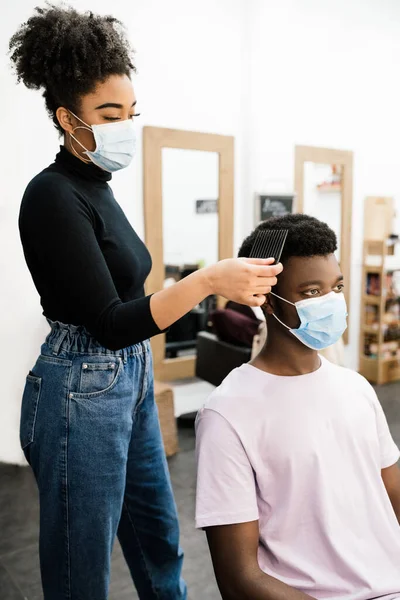 Afro-Amerikan güzellik kuaförü. Kendini koronavirüs salgınından korumak için hem maske hem de eldiven takan Afro-Amerikan bir müşteriyi soyup tarıyor.