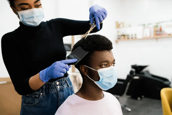 Afro-Amerikan güzellik kuaförü. Kendini koronavirüs salgınından korumak için hem maske hem de eldiven takan Afro-Amerikan bir müşteriyi soyup tarıyor.