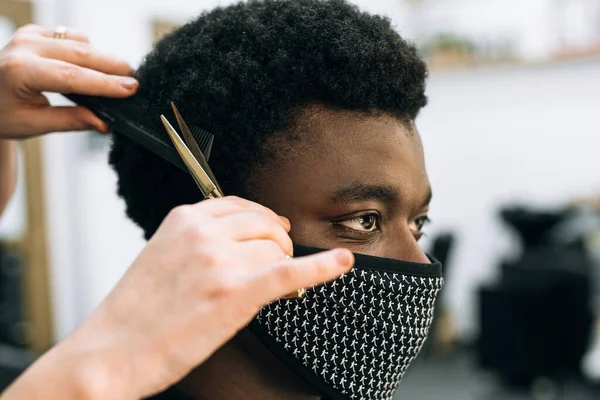 黒い男がコロナウイルスの顔に黒いマスクをつけて美容室で散髪してる 髪はアフロのようです ハサミは金だ — ストック写真