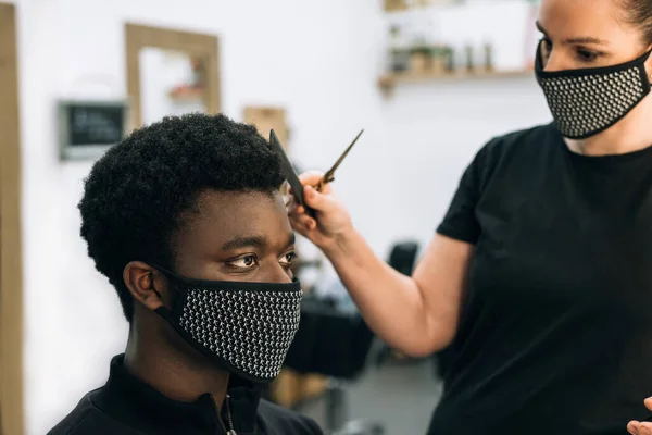 face of a black guy getting a haircut in a hair salon with a black mask on his face from the coronavirus. The hairdresser also wears a mask. The hair has it like the afro