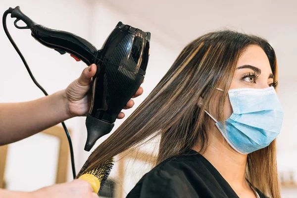 Saç kurutma makinesi taşıyan bir kuaförün eli Coronavirus yüzünden beyaz maskeli güzel bir kızın saçını kurutuyor. Kızın uzun saçları var.