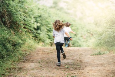 İki kardeş 4 yaşında kahverengi bir oğlan ve 7 yaşında sarışın bir kız çok güzel yeşil ağaçların ve sarı güneş ışığının arasından kameraya doğru koşuyorlar.
