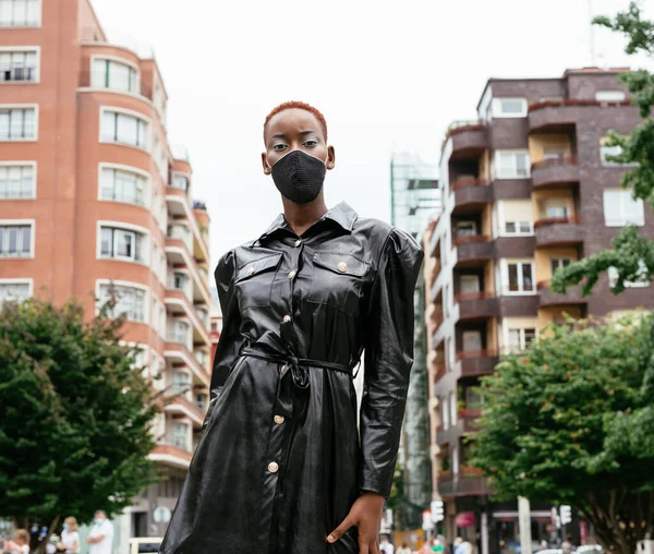 Beautiful black girl model with mask due to the coronavirus pandemic covid 19 walking in style on the street with a nice black dress