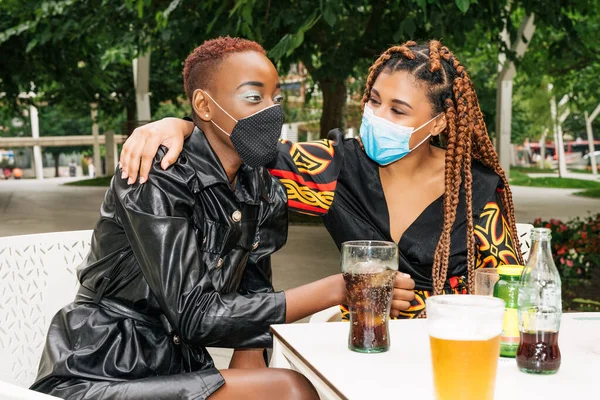 İki çok ırklı, siyah ve melez arkadaş bir bar terasında içiyorlar ve Coronavirus Codemic 19 salgını yüzünden yüz maskeleriyle birbirlerine sarılıyorlar.
