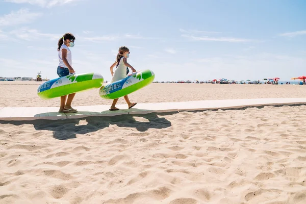 Yüzü maskeli ve gözlüklü iki kız kollarında renkli şişirilebilir bir çark ile kumsala doğru yürüyorlar. Covid 19 Coronavirüs salgınının ortasında tatilin tadını çıkarıyorlar.