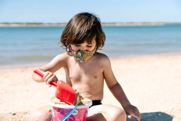 Child with face mask on vacation playing and enjoying toys on the sand of a beach next to the sea water in the middle of the coronavirus pandemic covid19