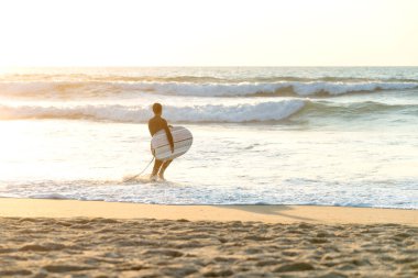 Kollarında sörf tahtası olan sörfçü gün batımında denize giriyor.