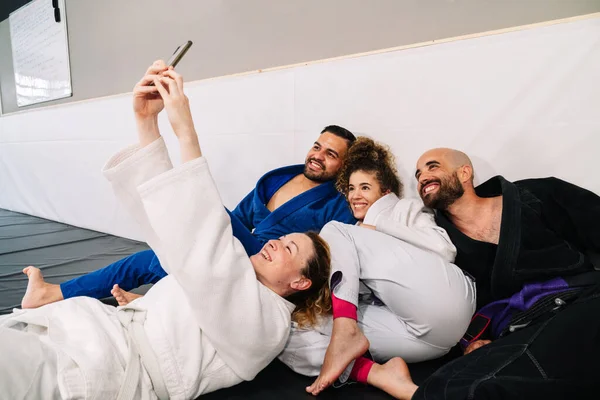 Portrait of four companions of martial arts like judo using kimono sitting on the carpet of the floor and leaning on the wall of the gym, making a selfie with the mobile very happy after a training