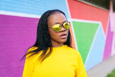 Yansıtıcı gözlüklü şık afro kadın, renkli şehir arka planı.