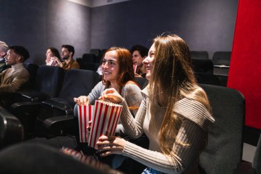 Genç kadınlar, sıcak bir sinemada birlikte film izlerken gülüyor ve patlamış mısırdan zevk alıyorlar, eğlence ve mutluluk anlarını paylaşıyorlar.