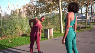 Kadın sporcular, doğal arka plana sahip kentsel park ortamında dinamik açık hava antrenmanı sırasında birbirlerini destekleyerek fitness antrenmanlarını senkronize ediyorlar.