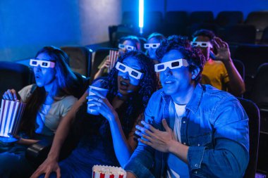 Üç boyutlu gözlük takan bir grup genç sinemada sürpriz bir film izliyor, patlamış mısır yiyor ve gazoz içiyor.