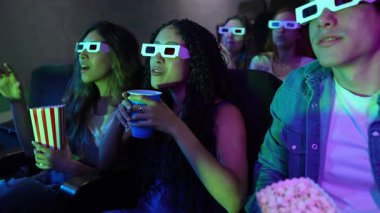 Genç arkadaşlar 3D gözlük takıyor, patlamış mısır yiyor ve yavaş çekimde sinemada film izlerken soda içiyorlar.