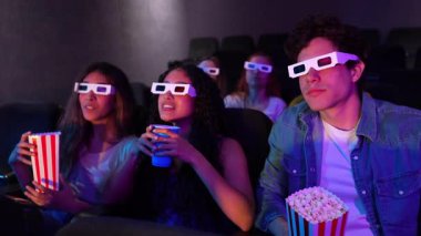Genç arkadaşlar 3D gözlük takıyor, patlamış mısır yiyor ve sinemada film izlerken soda içiyorlar.