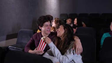 Genç çift film gösterimi sırasında neşeli anları paylaşıyor, gülüyor ve patlamış mısırın tadını çıkarırken loş ışıklı sinema salonunda yan yana oturuyorlar.