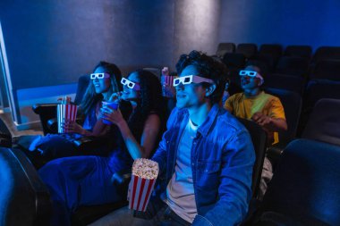 Üç boyutlu gözlük takan, sinemada film izleyen, patlamış mısır yiyen ve gazoz içen bir grup genç.