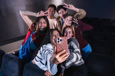 Bir grup neşeli çok ırklı üniversite öğrencisi konferans salonunda akıllı bir telefonla selfie çekiyor.