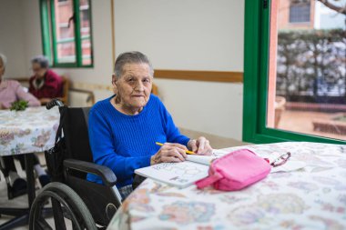 Huzurevindeki bir masada tekerlekli sandalyede oturan yaşlı bir kadın boş zamanlarında etkinlik yapıyor, refahı ve bakımı teşvik ediyor.