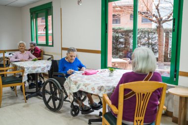 Üst düzey kadınlar ortak bir odada masalarda oturuyor, bir huzurevinde sosyal etkileşim ve boş zamanların keyfini çıkarıyorlar.