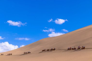 Turist grubu kum dağı Mingsha Shan çölünde deveye biniyor ya da Çin 'in Gansu kentindeki Dunhuang şehrinde İpek Yolu' nun bir parçası olarak kervanla şarkı söyleyen kum tepelerinde..