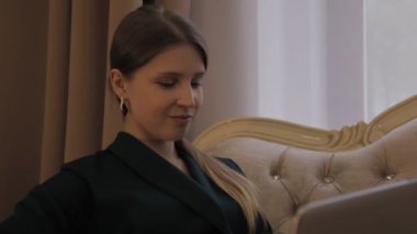Siyah gömlekli Slav kız, pencere kenarındaki kanepede dizüstü bilgisayarla oturuyor ve duygularını gösteriyor. Kadın ekrana bakar, pencereden bakar, gülümser, mutludur. Genç bayanın (yüzü) yakın plan
