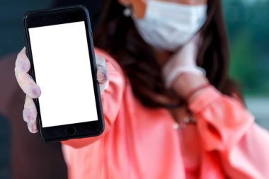Tıbbi maskeli bir kadın ve elinde telefon olan tek kullanımlık eldivenler. Koruyucu maskeli endişeli kadın sokakta boş akıllı telefon ekranı gösteriyor. Yazarın yeri. Yazı veya logo için arkaplan boşluğu
