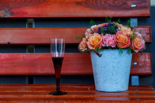 Flowers on bench. Bouquet of roses in a bucket stands on an old wooden bench next to glass of red wine in summer garden at dusk. Space for your creativity, space for an inscription or logo