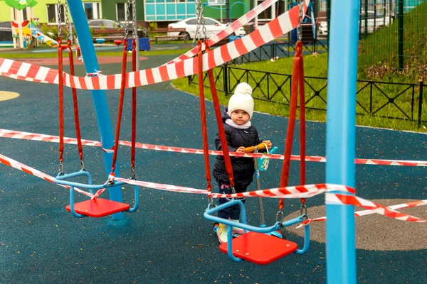 Child on closed Playground. Sad upset girl on scooter on closed outdoor area. The Playground is closed with warning tape. Young girl looks at closed Playground during Covid 19 pandemic