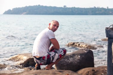 Yazlık elbiseli bir adam kayanın üzerinde oturuyor ve arka planda tropikal bir deniz var. Spor figürlü bir adam kameraya döndü. Seyahatler. Yaz yaşam tarzı, manzarası olan tropikal bir ülkede.