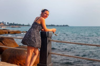 Günbatımında tropikal denizin arka planında bir kız. Yazlık elbiseli kadın çitte duruyor. Iskaladım. Kayıp seyahat. Rüya gören bir kadın. Yaz yaşam tarzı, arka planda balkon ve deniz ile plajda